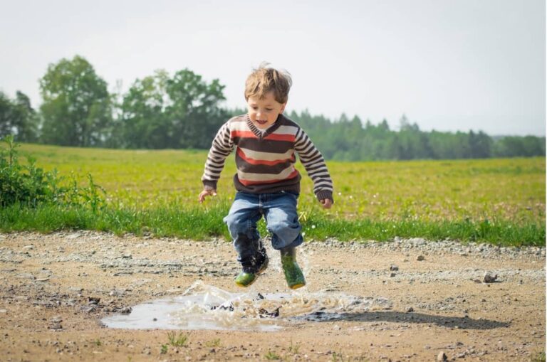 Chłopiec w pasiastym sweterku skacze po kałużach, w tle - wiejski krajobraz