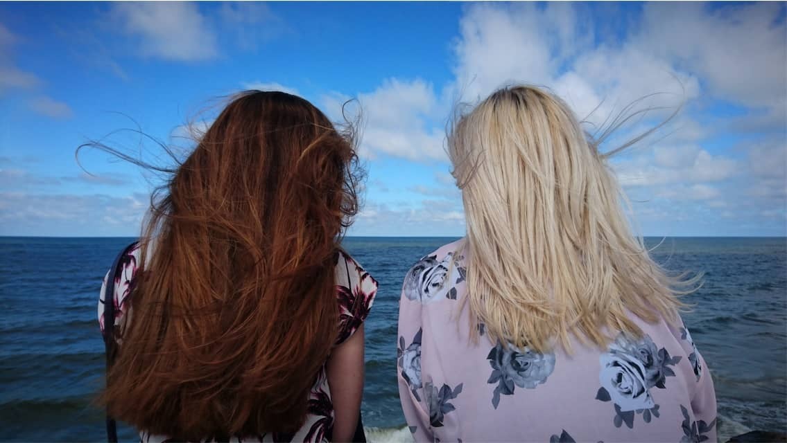 2 dziewczyny odwrócone tyłem nad brzegiem morza. Jedna jasnowłosa, druga rudowłosa