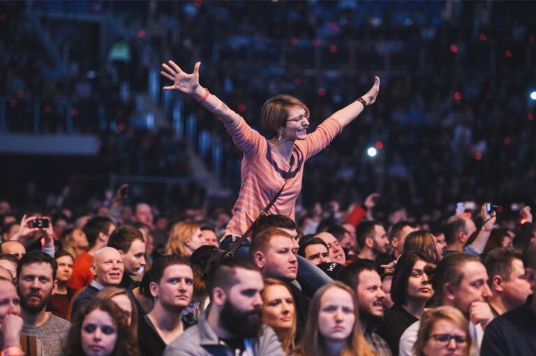 Tłum podczas koncertu. Kobieta w okularach i różowej bluzce rozkłada ręce, podniesiona do góry przez tłum
