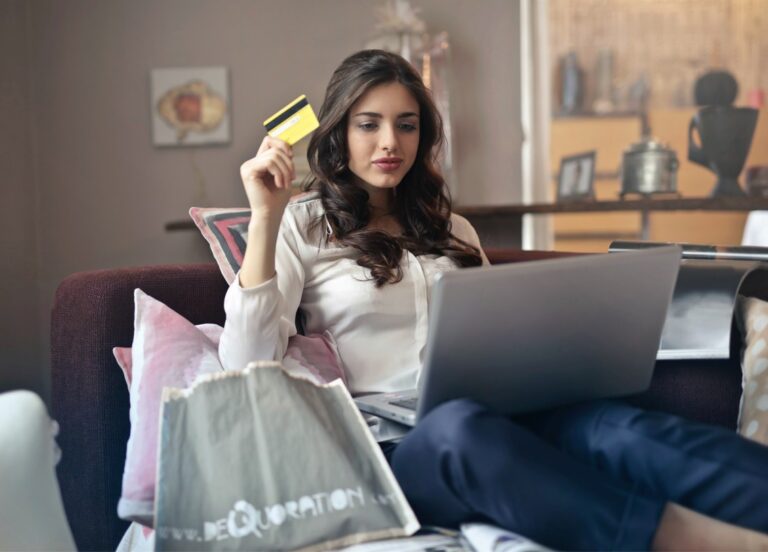 Długowłosa dziewczyna siedzi z laptopem na kolanach, w ręku trzyma kartę płatniczą, wokół niej - torby z zakupami