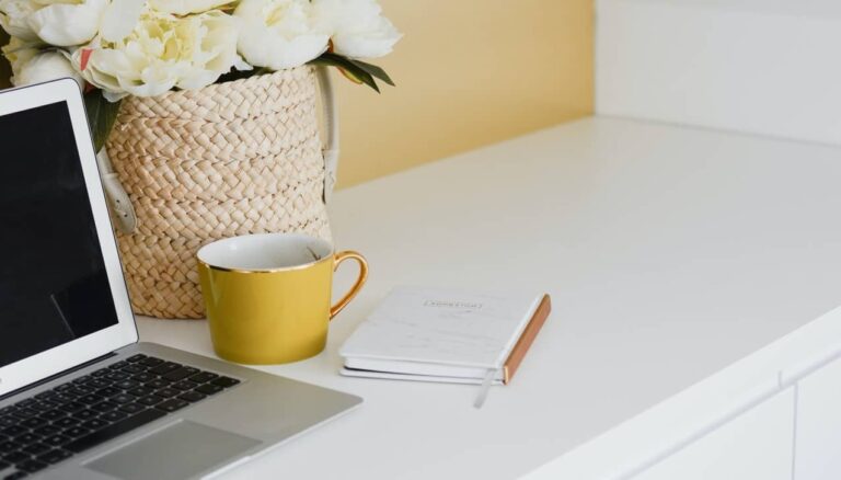 Na białym biurku leżą: laptop, żółty kubek, notes i kosz z kwiatami